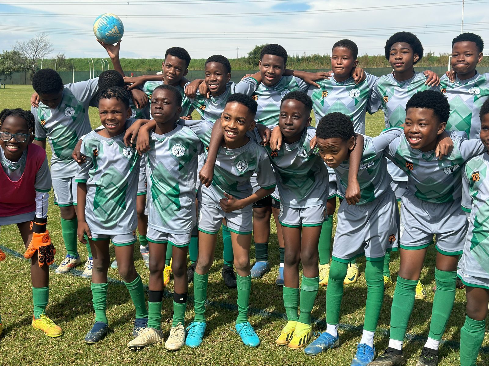 A youth soccer team in matching grey and green uniforms poses together on a grassy field, with one player holding a ball.