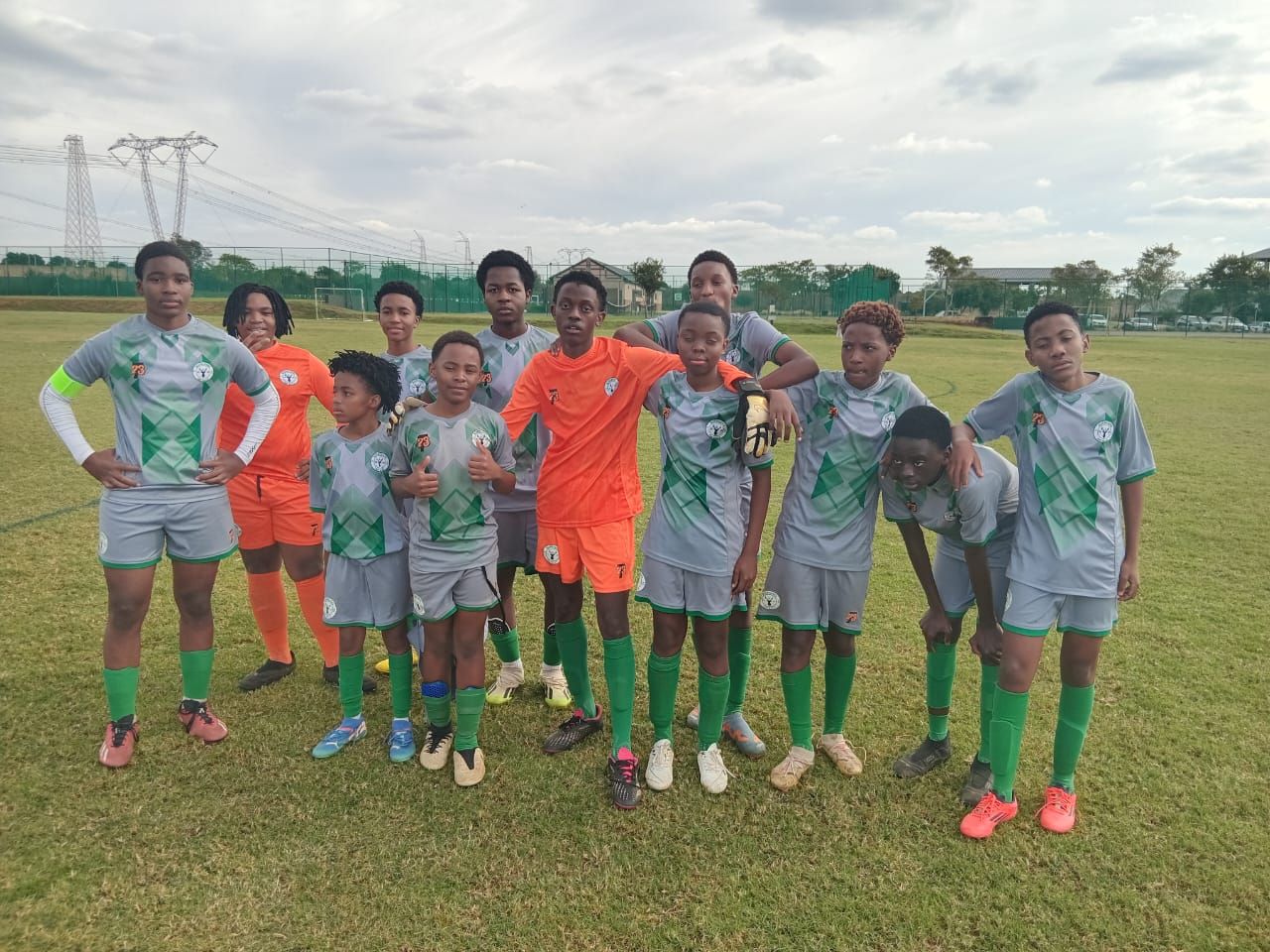 A group of young soccer players wearing grey and orange jerseys standing on a grass field under a cloudy sky.