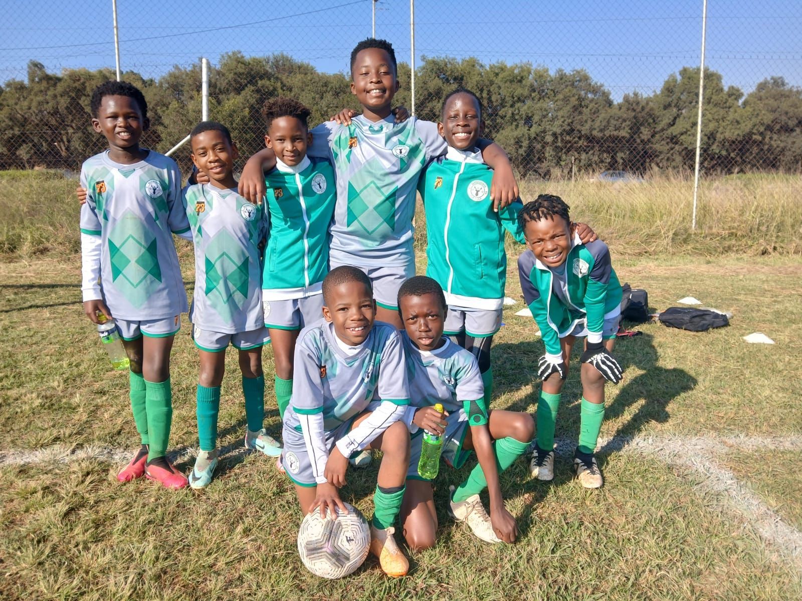 A youth soccer team in green and grey uniforms poses together on a grassy field, with one child holding a soccer ball.