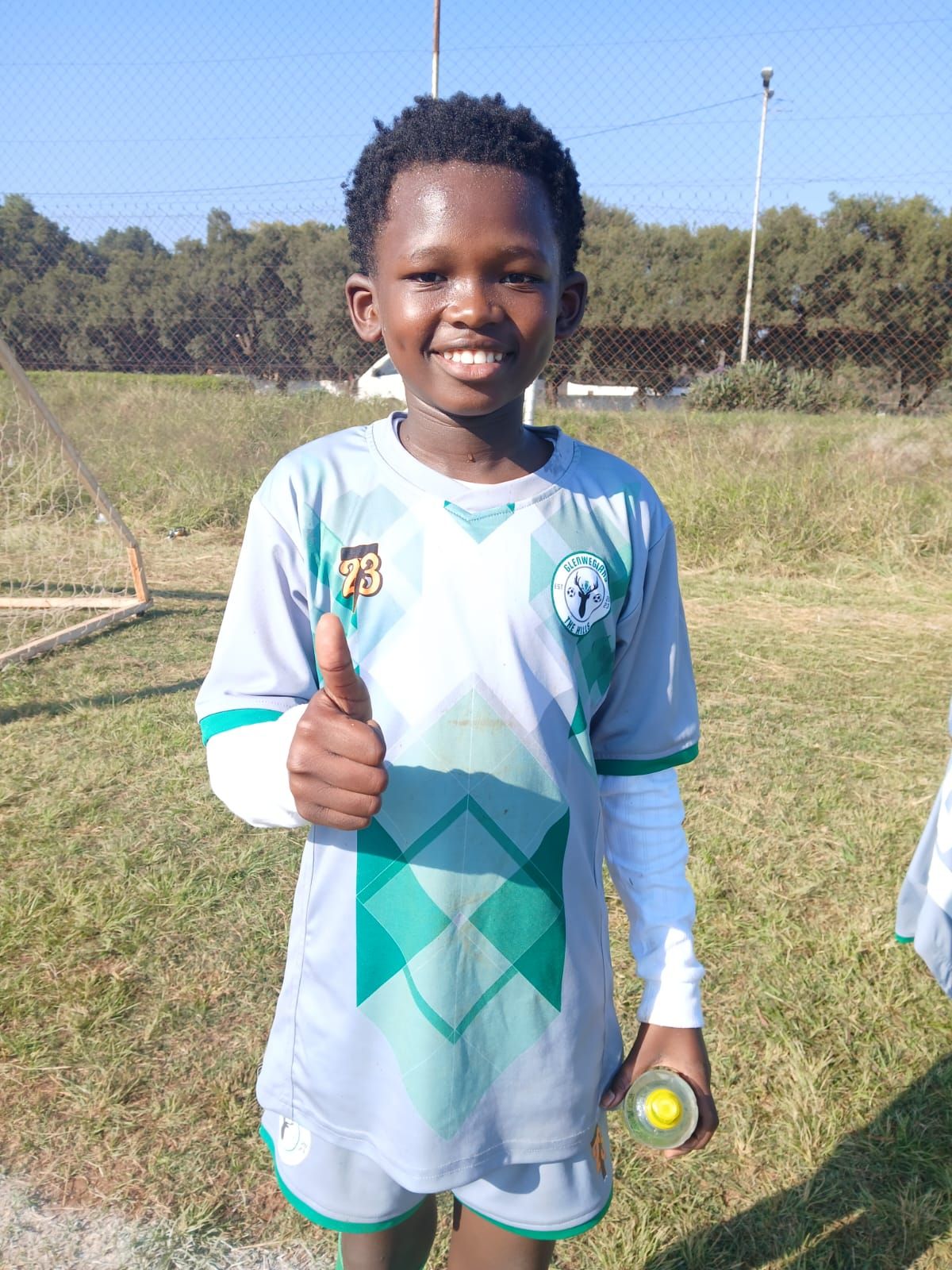 A smiling child in a grey and green soccer uniform gives a thumbs up while standing on a grassy field.