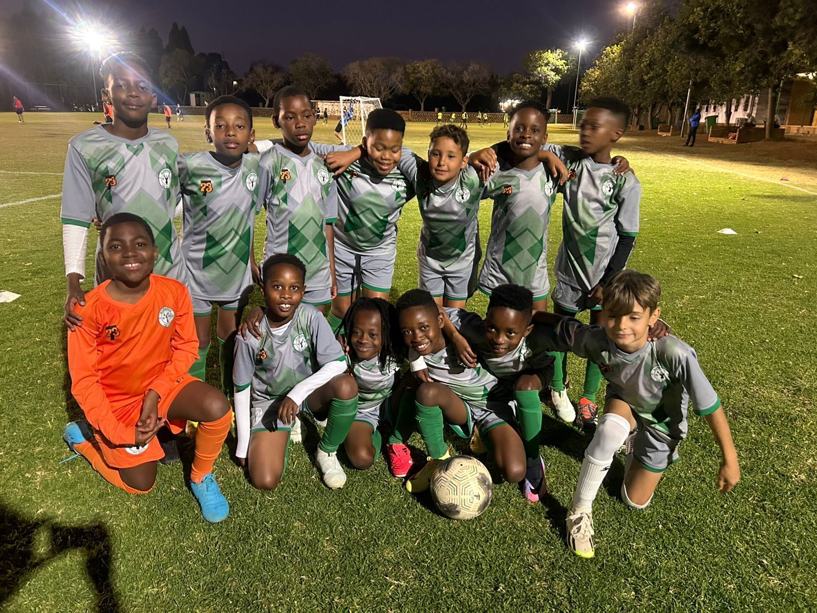 A youth soccer team in green and grey uniforms poses on a grass field under stadium lights with a ball at their feet.