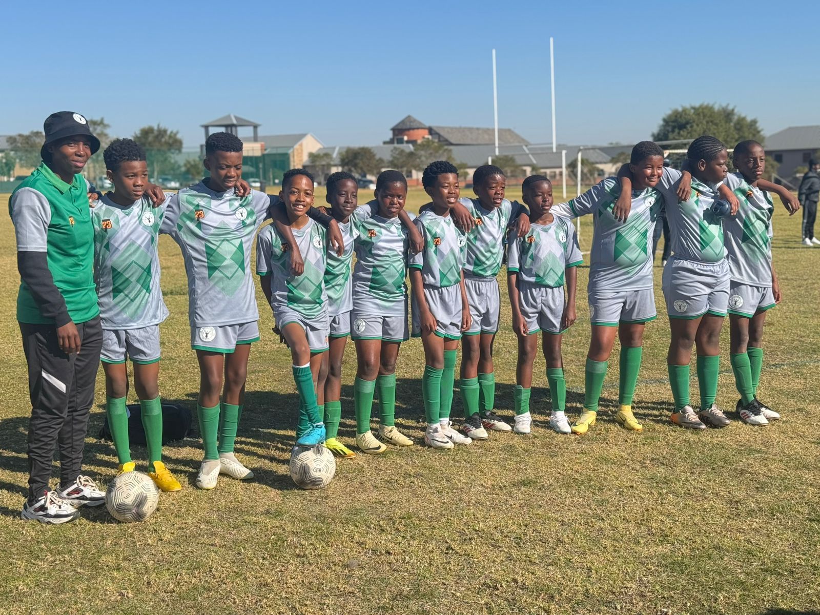 A youth soccer team in green and grey uniforms poses for a photo on a grassy field, with a coach standing to the left.