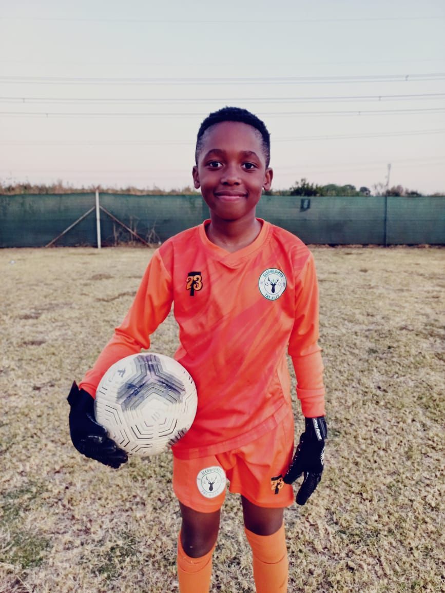 A soccer goalkeeper in an orange uniform holds a ball on a grass field under a clear sky.