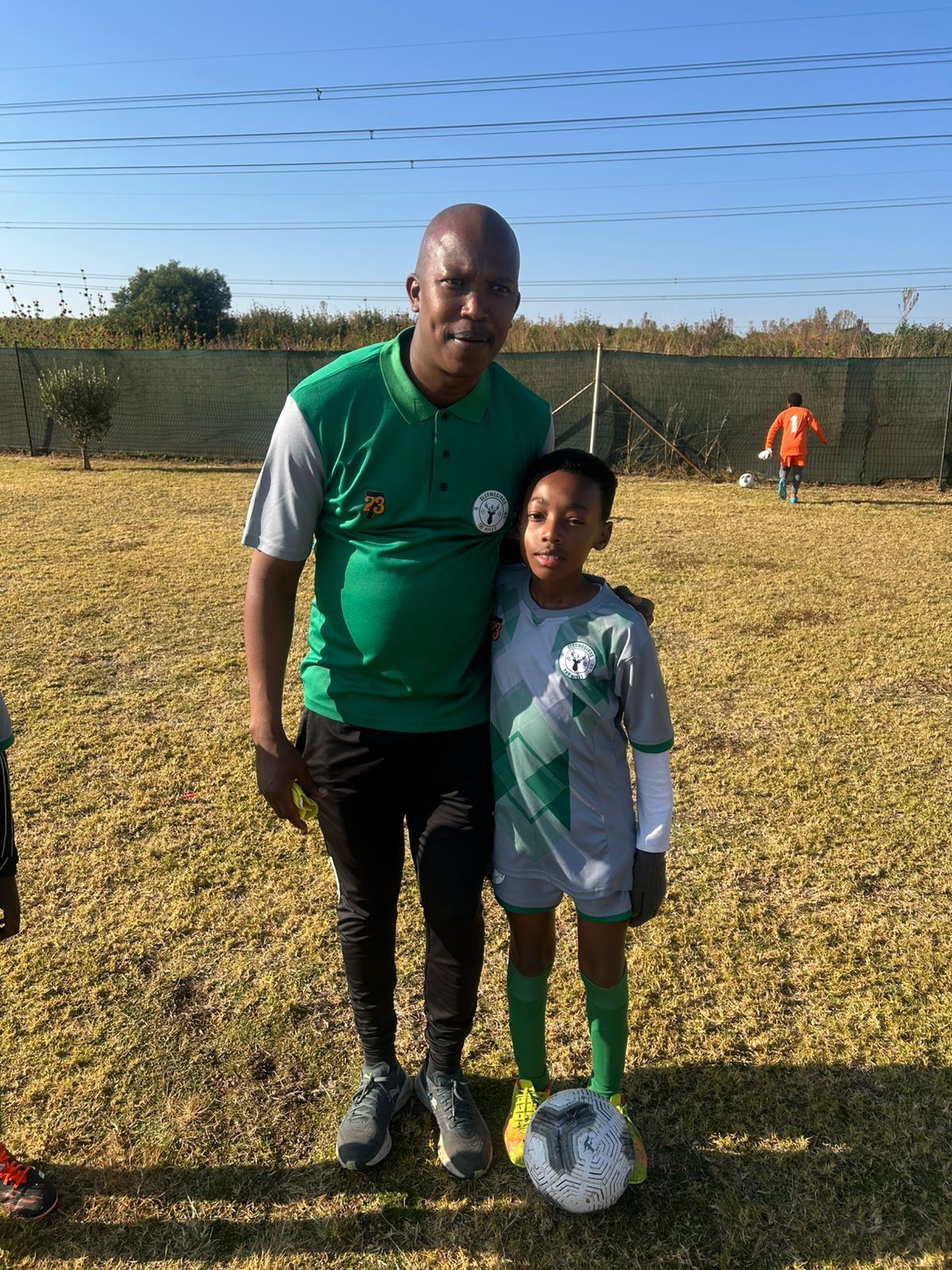 An adult and a child in soccer uniforms stand together on a grassy field, with a soccer ball at the child's feet.
