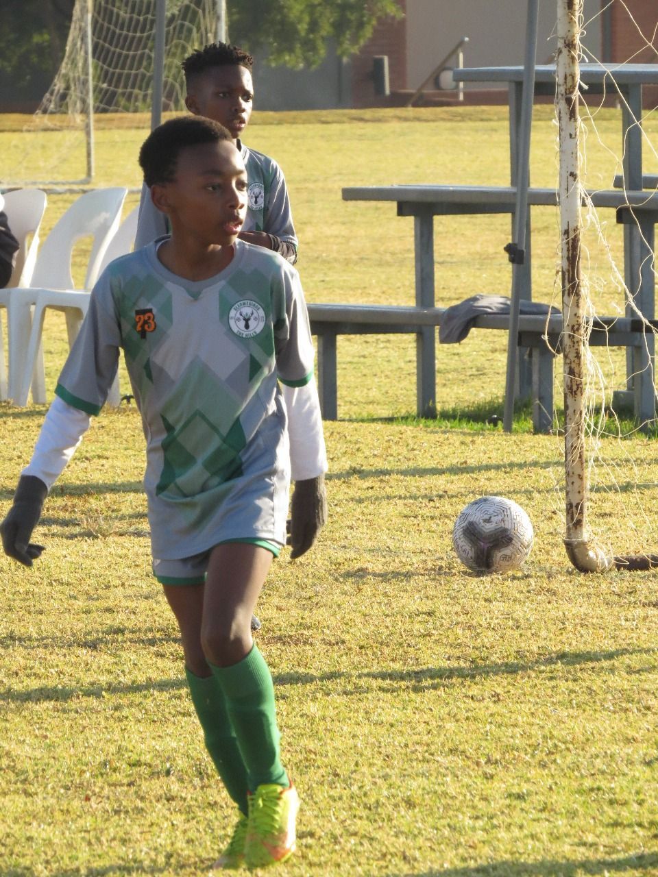 A youth soccer player in a grey jersey and green socks walks on a grass field near a goal net and a soccer ball.