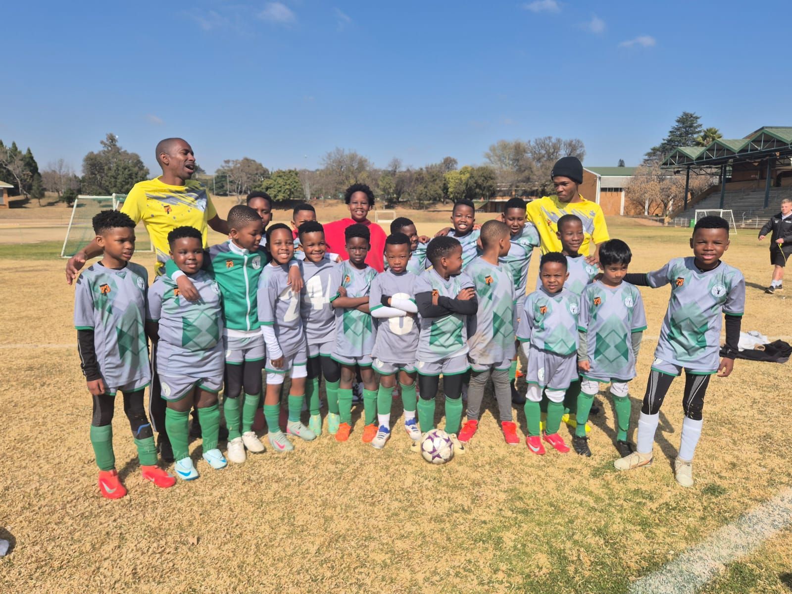A youth soccer team in grey and green uniforms poses with two coaches on a dry, grassy field under a clear blue sky.