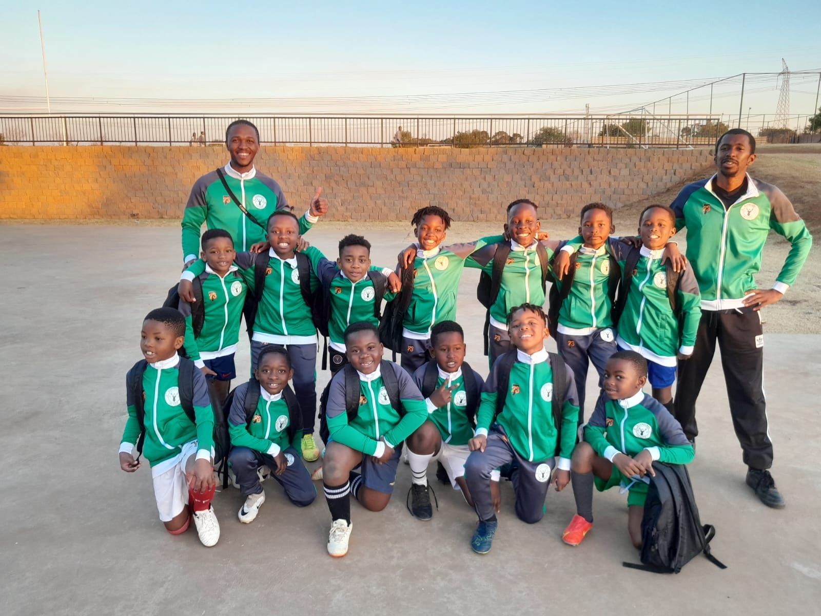 A soccer team and two coaches in matching green and black uniforms pose together on a paved field.