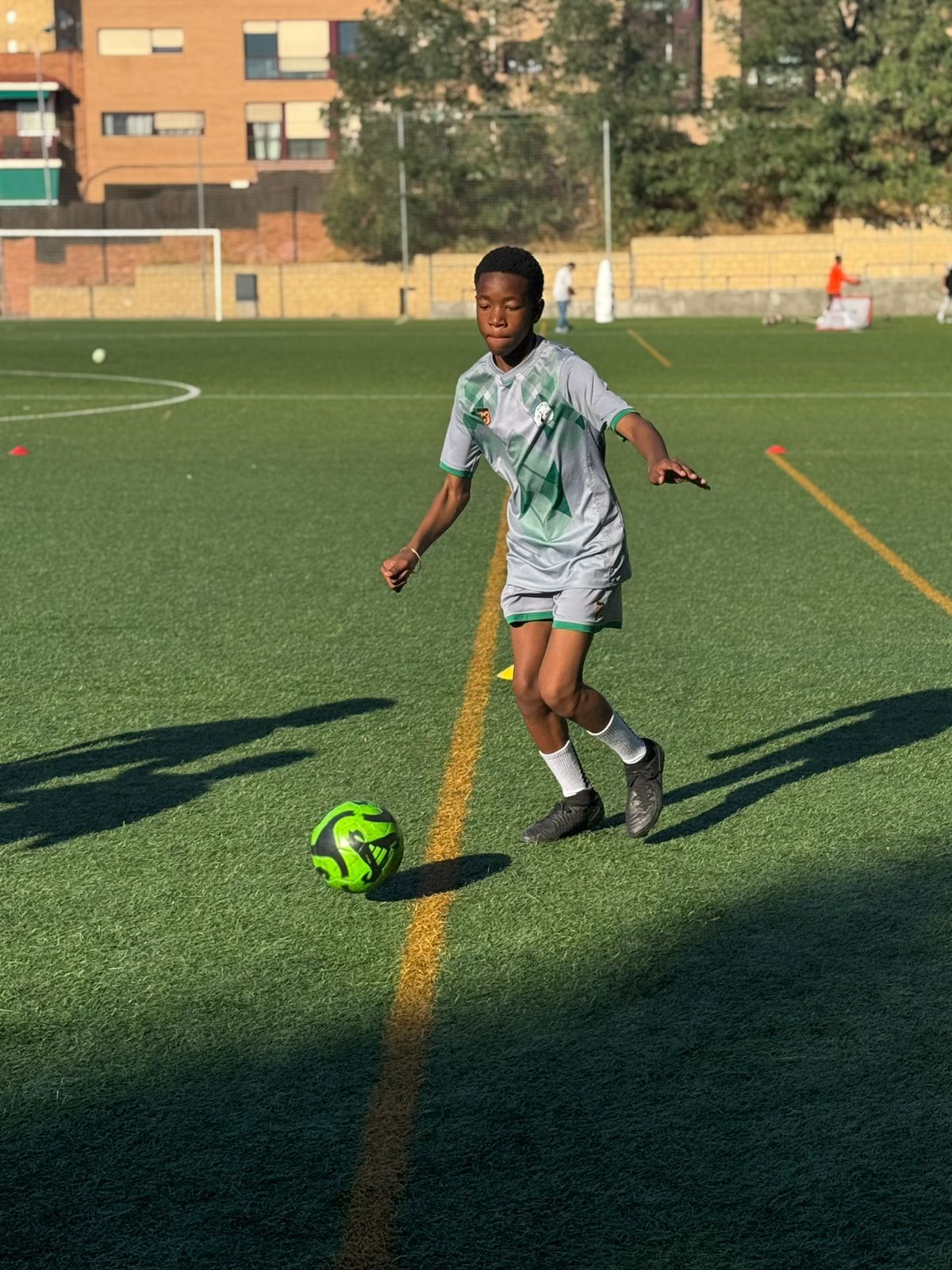 A person in a light grey soccer uniform dribbling a neon green ball on an outdoor synthetic field.