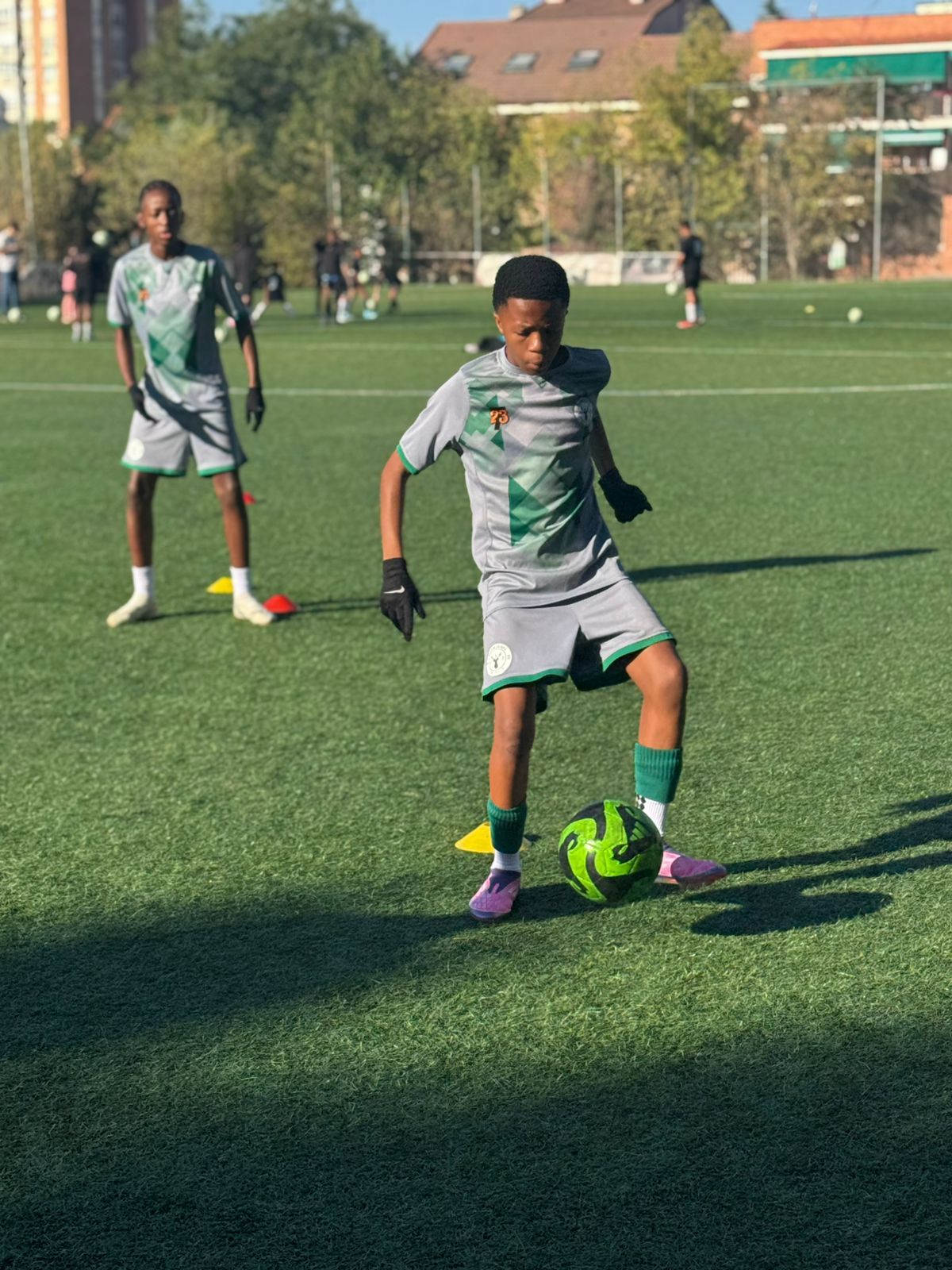 Two soccer players in gray and green kits practice on a sunny artificial turf field with cones and a soccer ball.