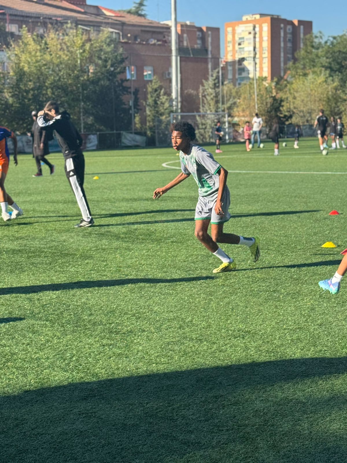 A soccer player in a green and white uniform jogs across a sunny artificial turf field during a training session.
