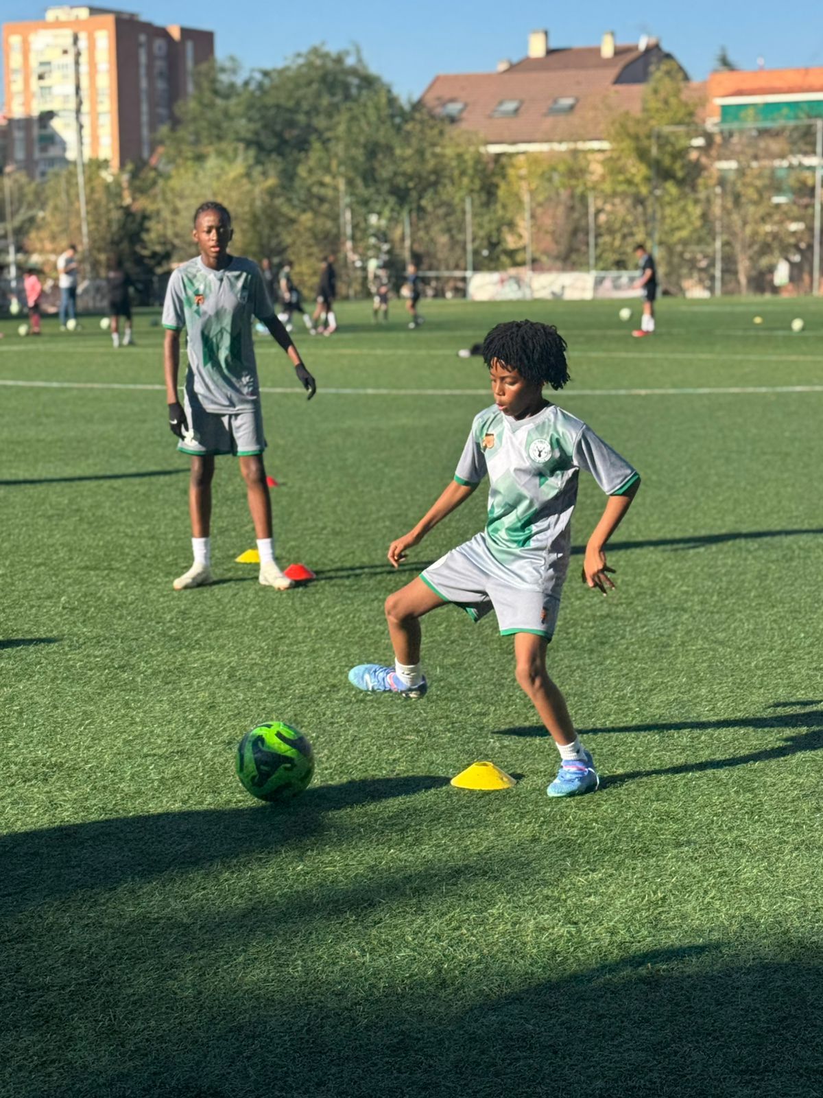 Two youth soccer players in gray uniforms practice drills on a sunny artificial turf field with cones and a soccer ball.