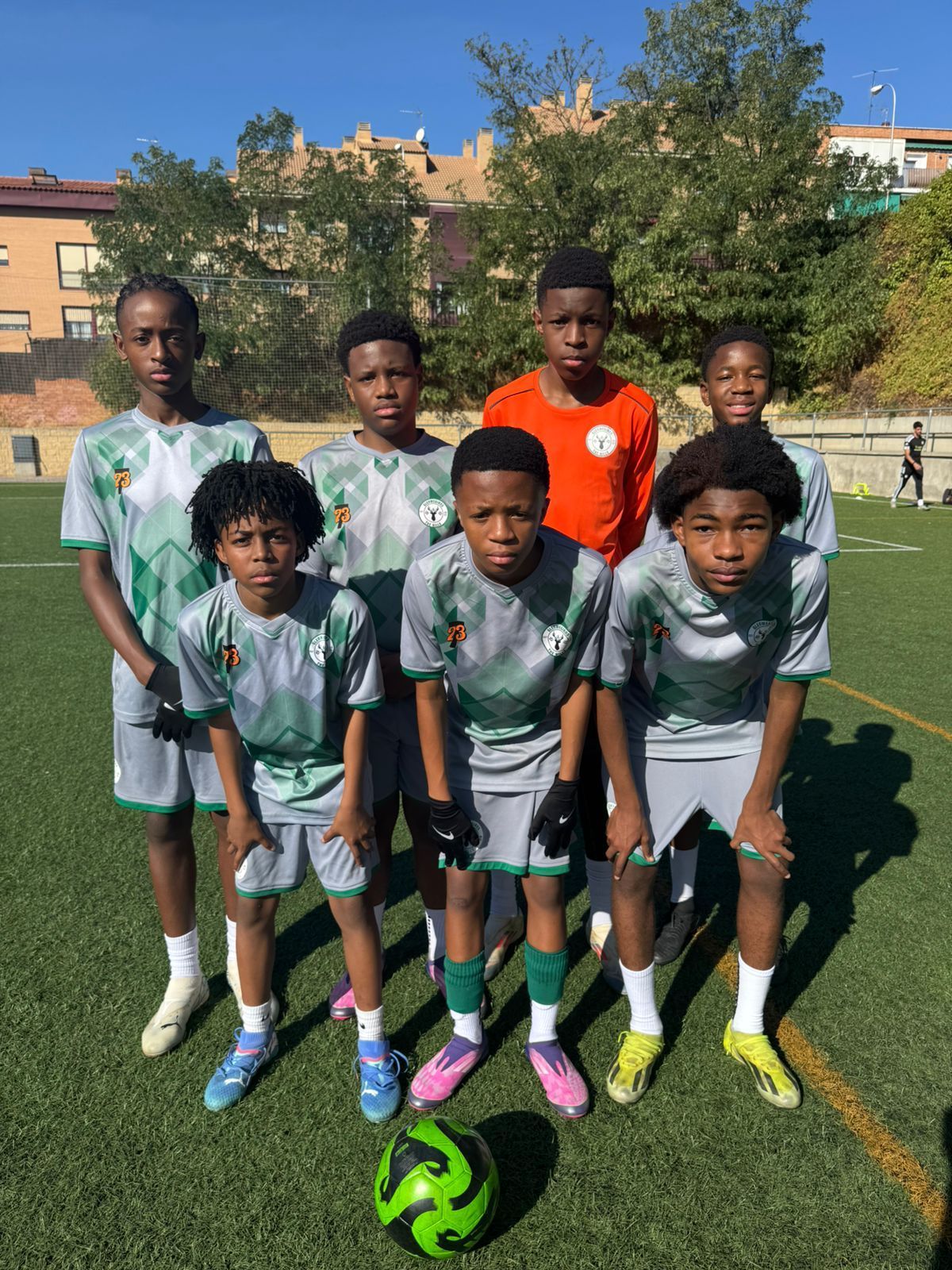 A youth soccer team poses for a photo on a green field, wearing grey patterned jerseys, with a soccer ball in front.