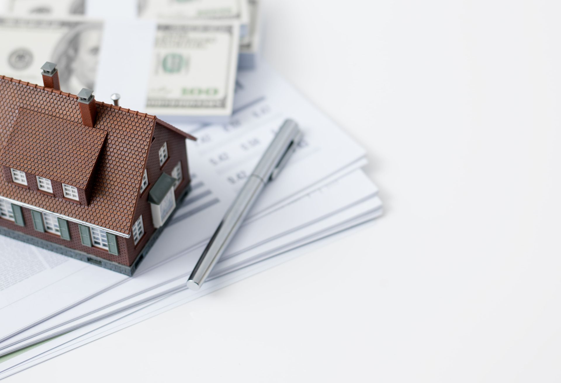 A model house is sitting on top of a pile of papers next to a pen and money.