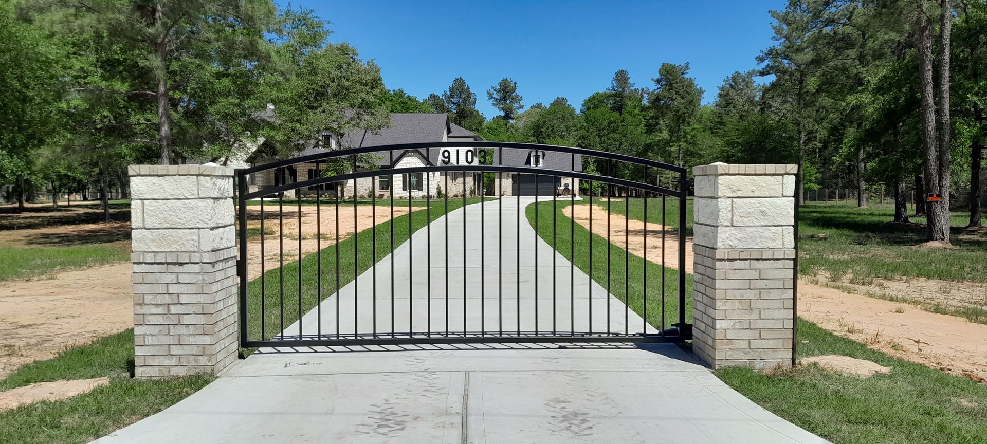 A black metal gate set between two stone pillars, opening to a concrete driveway leading to a house in a wooded area.