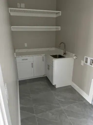 Empty laundry room with sink, white cabinets, wall shelves, and gray tile floor.
