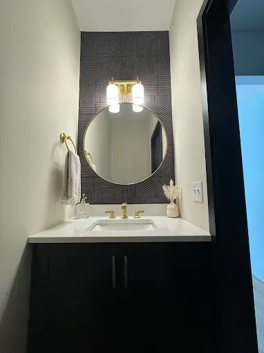 Small bathroom vanity with round mirror, brass light, black cabinet, and white countertop.