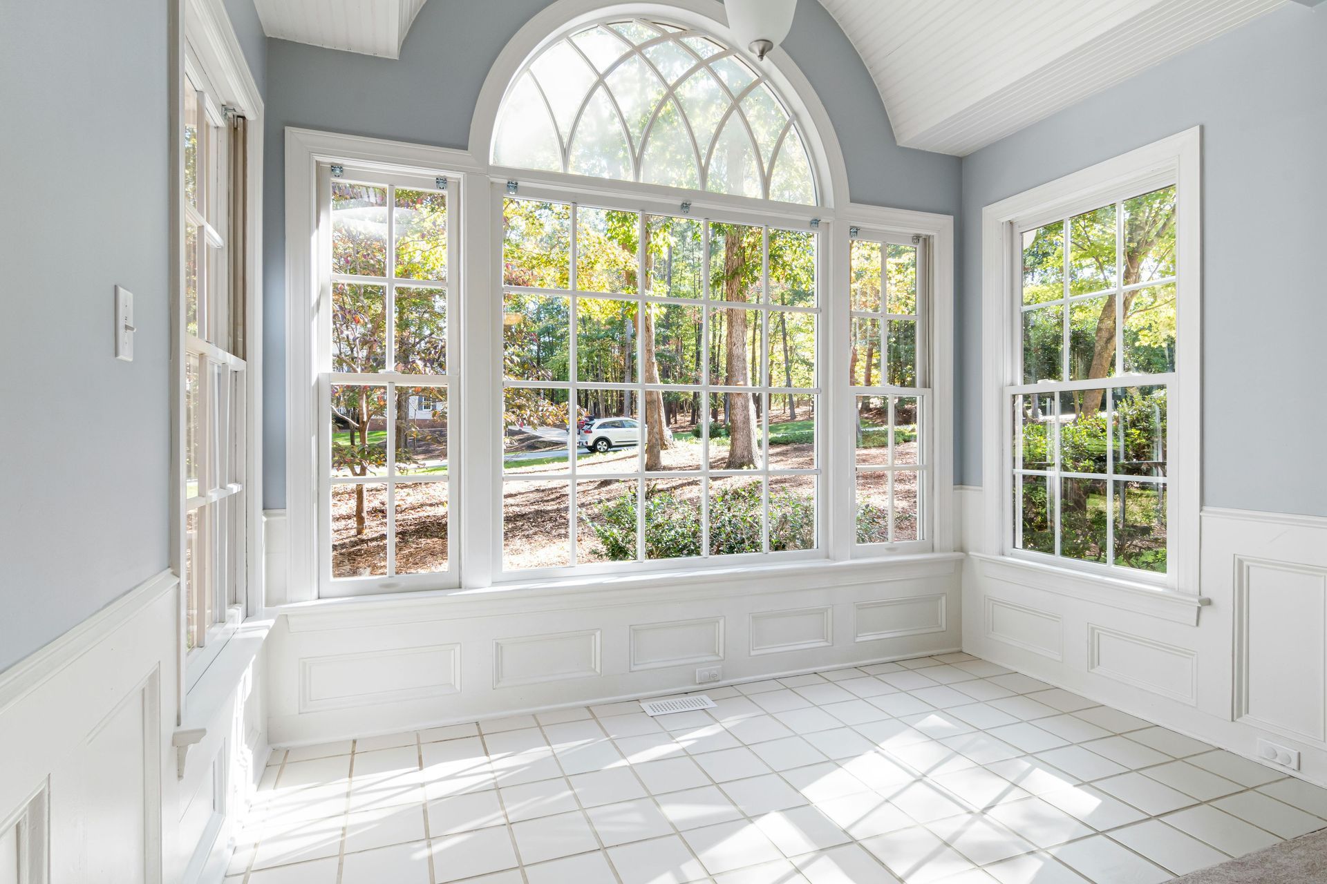 Bright sunroom with arched windows, white paneled walls, and a tiled floor overlooking a garden.