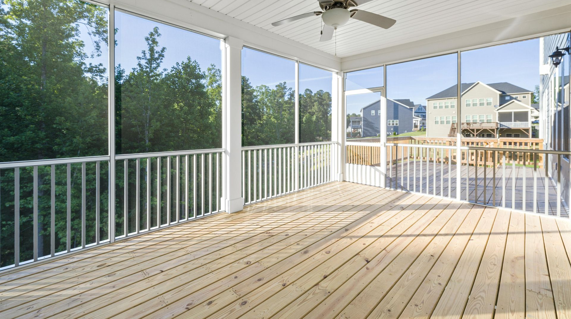 Spacious covered wooden deck with white railings, ceiling fan, and view of trees and nearby houses