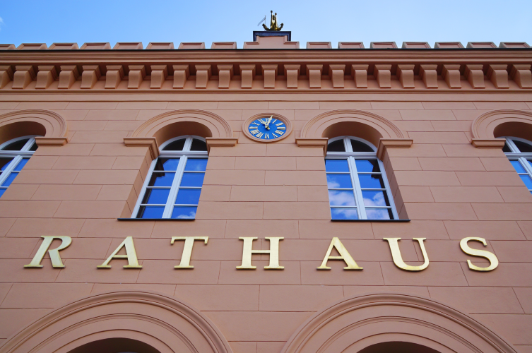 Fassade eines roten Backstein-Rathauses mit Bogenfenstern und goldener Schrift vor blauem Himmel
