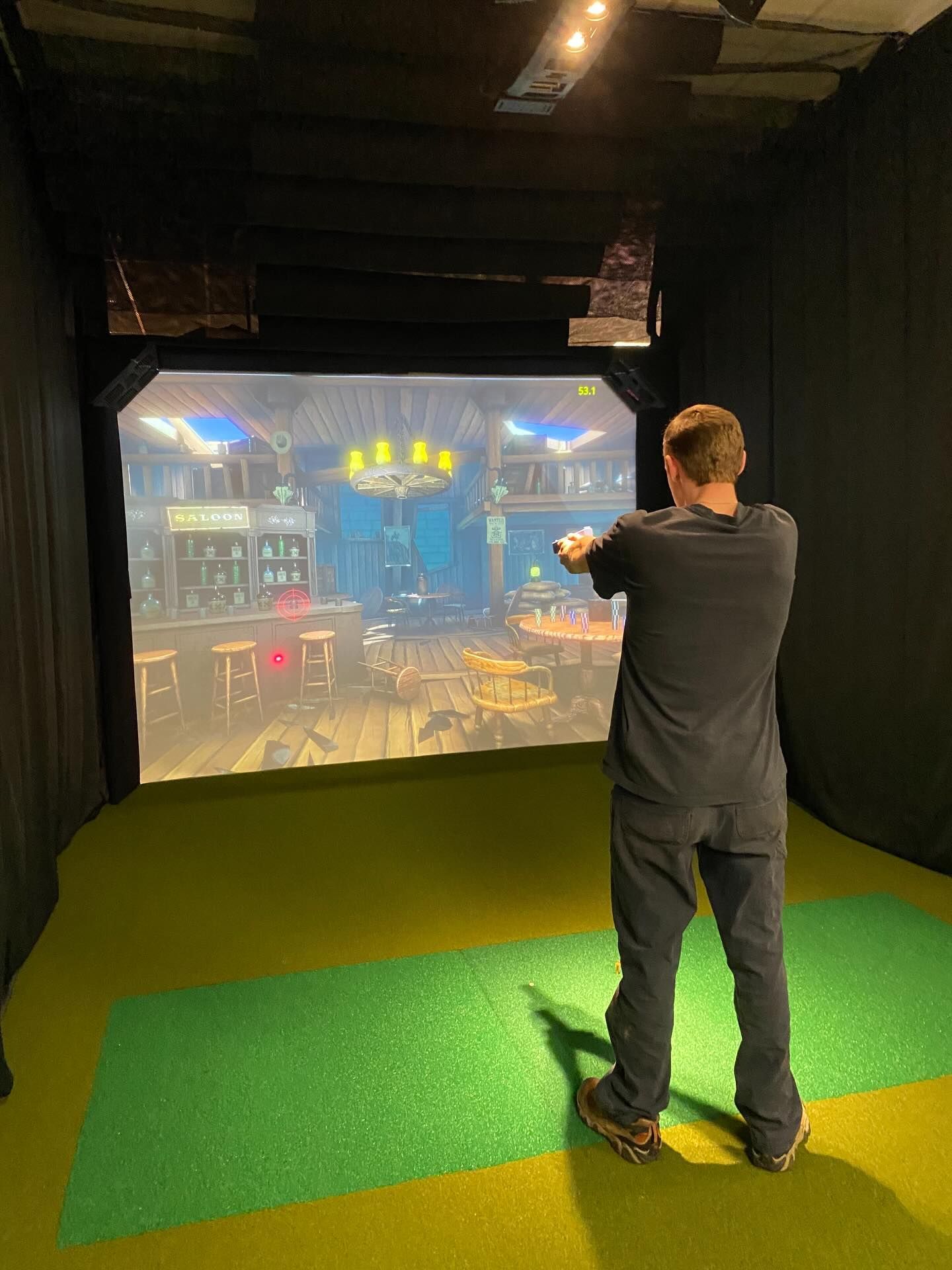 Person aiming a controller in a dark VR gaming room with a projected game scene on the wall