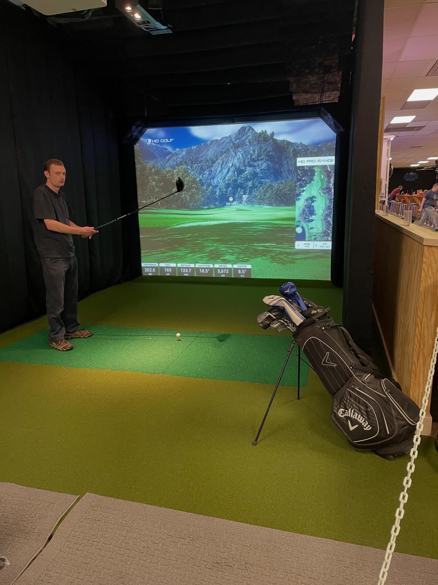 Man in golf simulator bay hitting a ball toward a projected course screen, with clubs in a bag beside him.