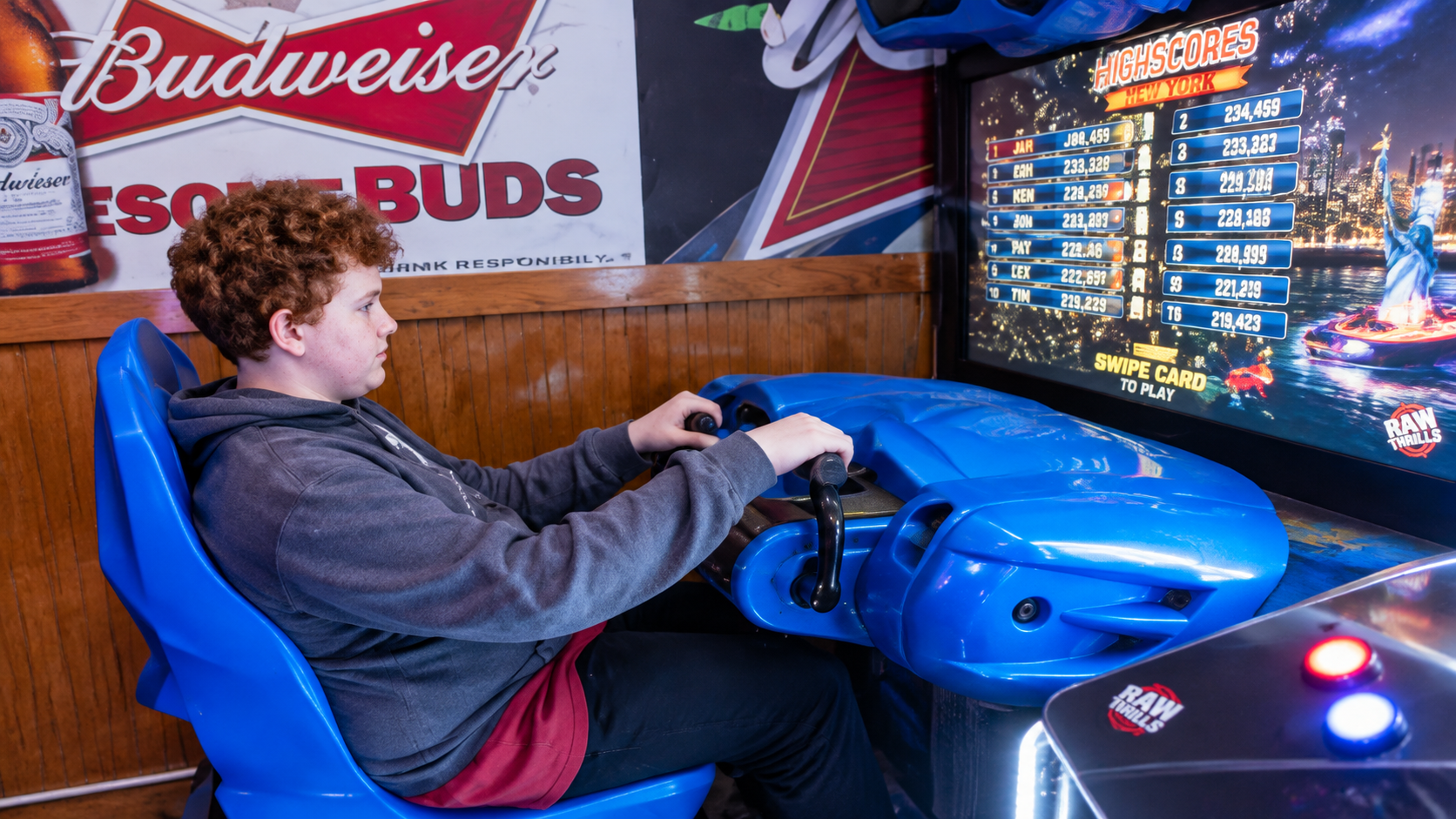 Person seated in a blue arcade racing game, steering with a focused expression, with a Budweiser sign in the background