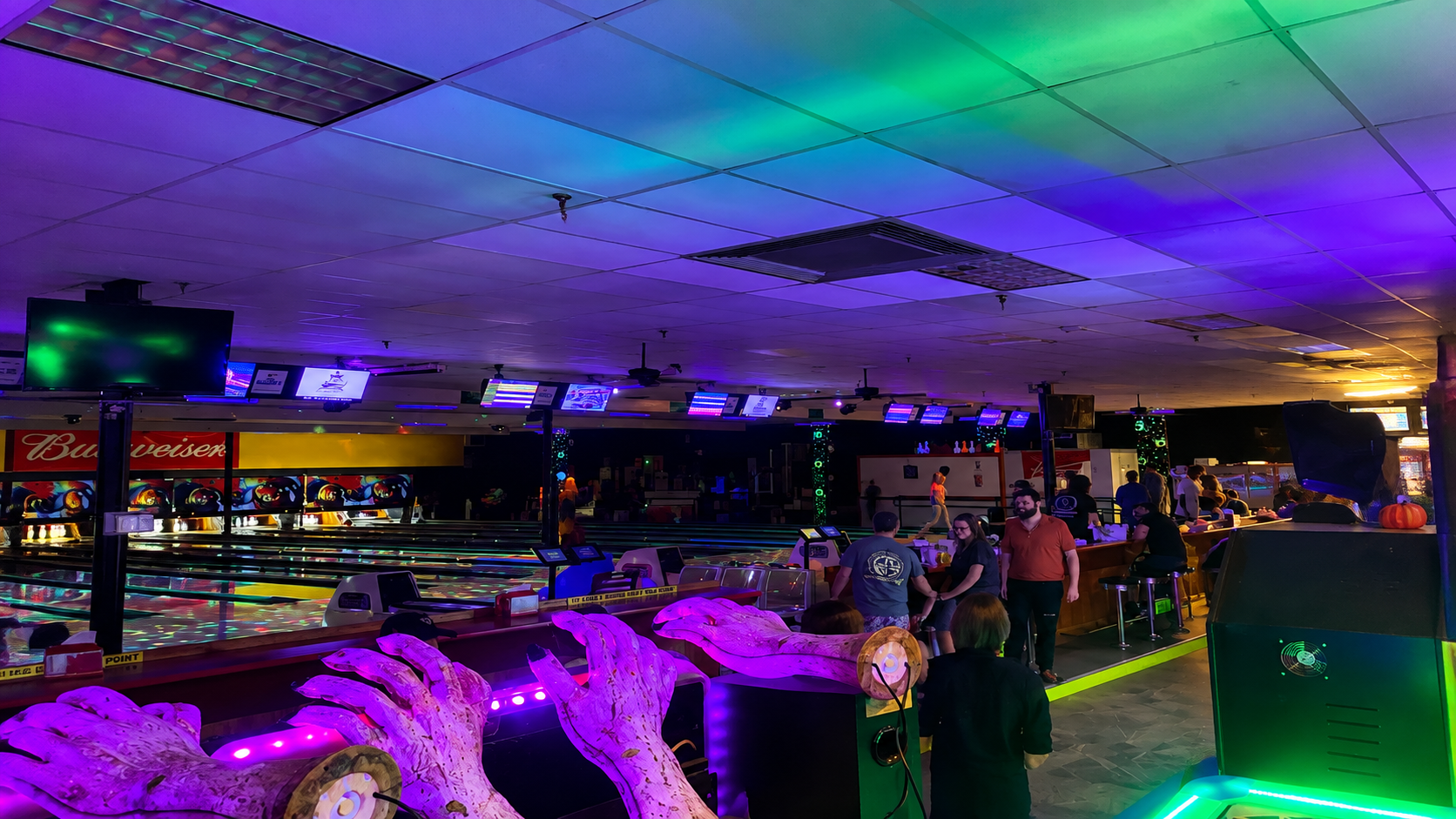 Neon-lit bar interior with purple and green lights, ceiling panels, and patrons standing near the counter