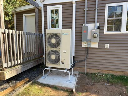 Outdoor heat pump unit beside a house, with two fans mounted on a concrete pad near utility meters.