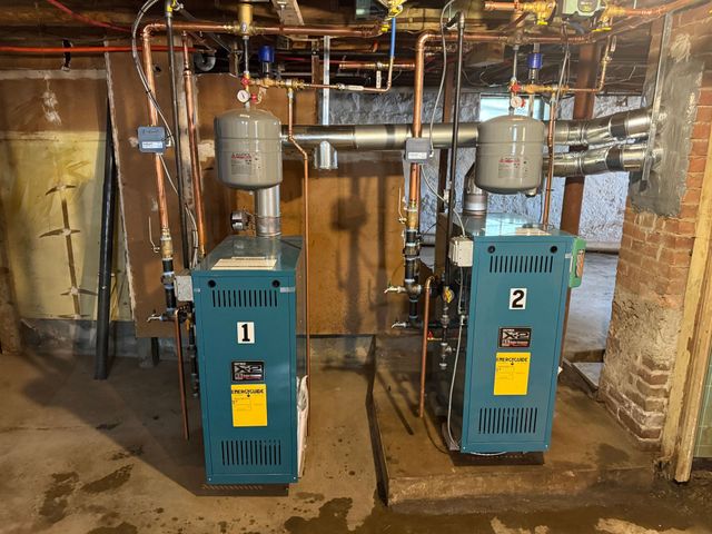 Two blue industrial boilers in a basement utility room with pipes and brick walls.