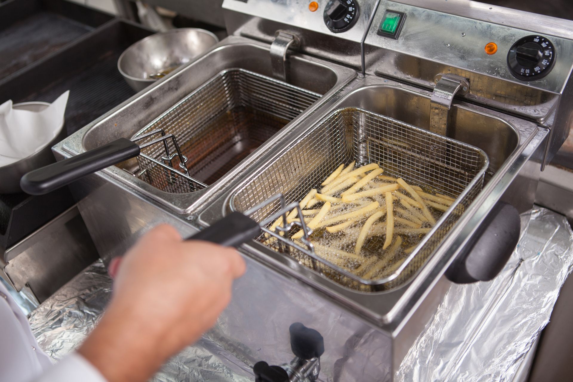 Manos friendo papas fritas en freidoras de acero inoxidable en una cocina comercial.