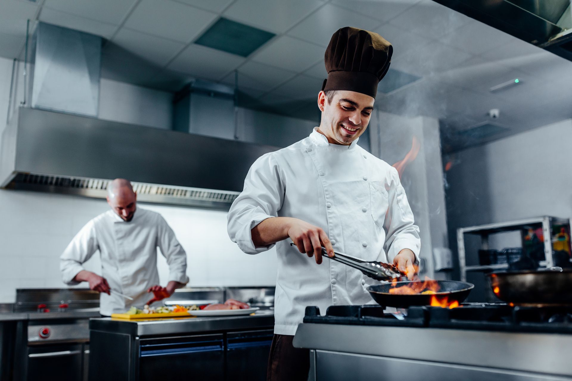 Los chefs cocinan en una cocina comercial de acero inoxidable, uno revolviendo una sartén en llamas y otro preparando la comida en la parte trasera.