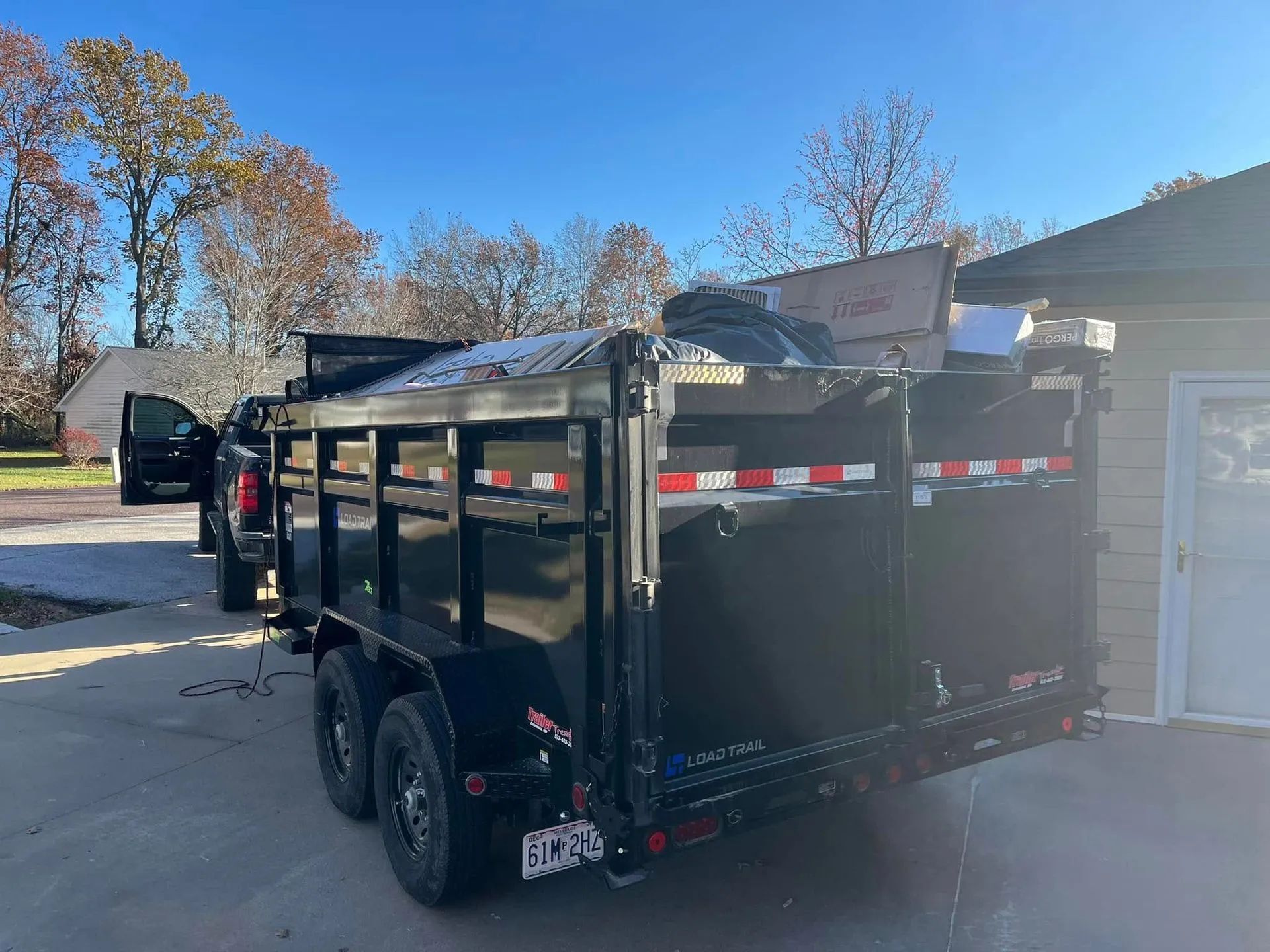 Black utility trailer with side rails parked in a driveway beside a house and trees.