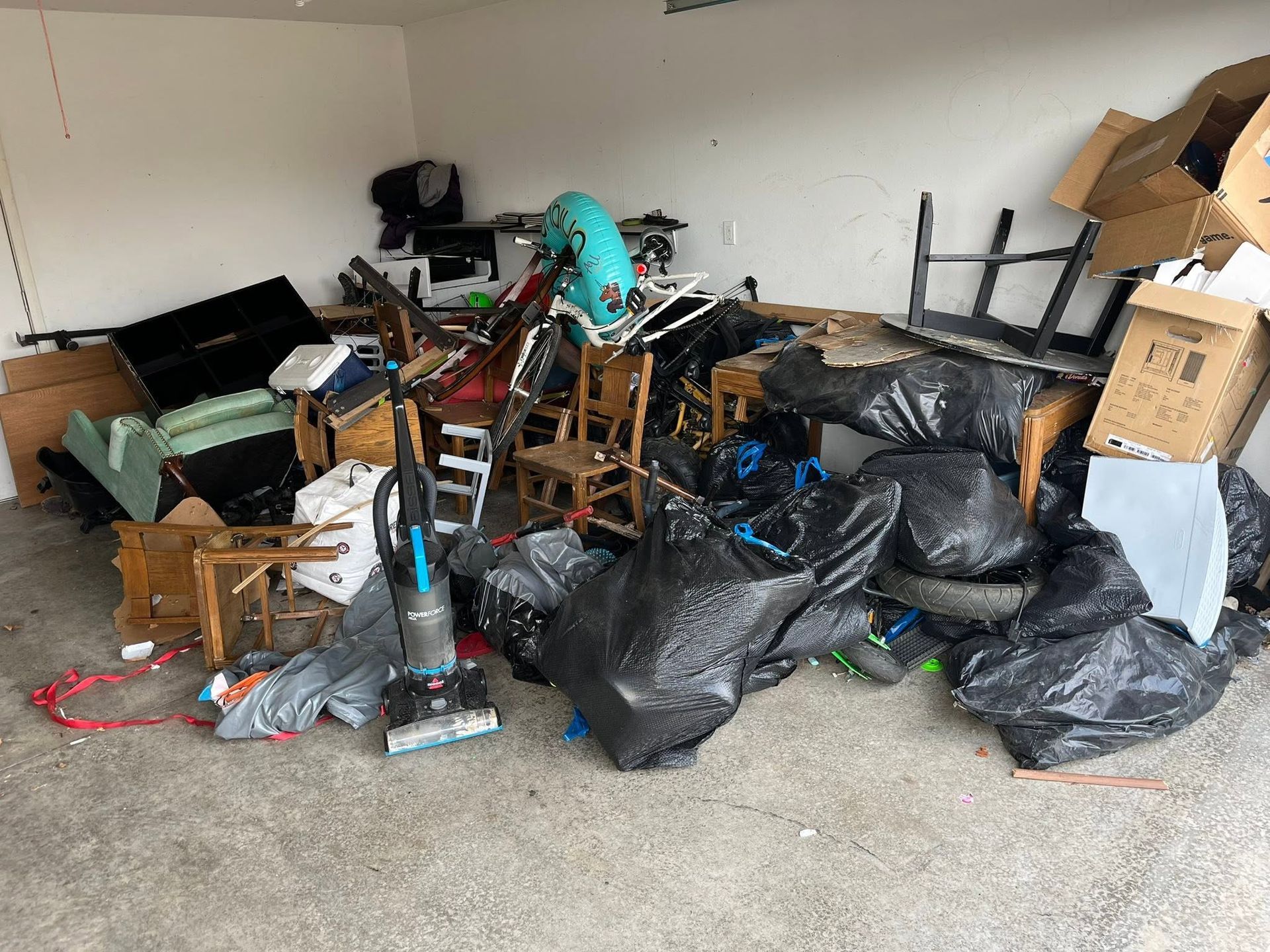 Cluttered garage with piled furniture, boxes, tools, and garbage bags scattered across the floor.