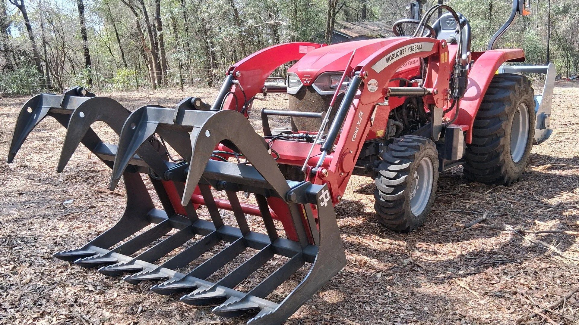 Red tractor with front grapple attachment parked on a leaf-covered dirt trail in a wooded area