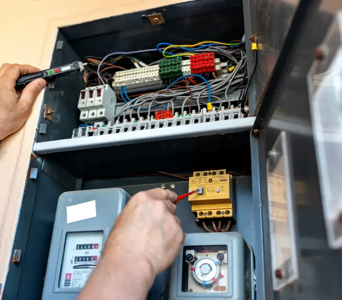 Hands testing live electrical components inside an open junction box with a multimeter and voltage tester.