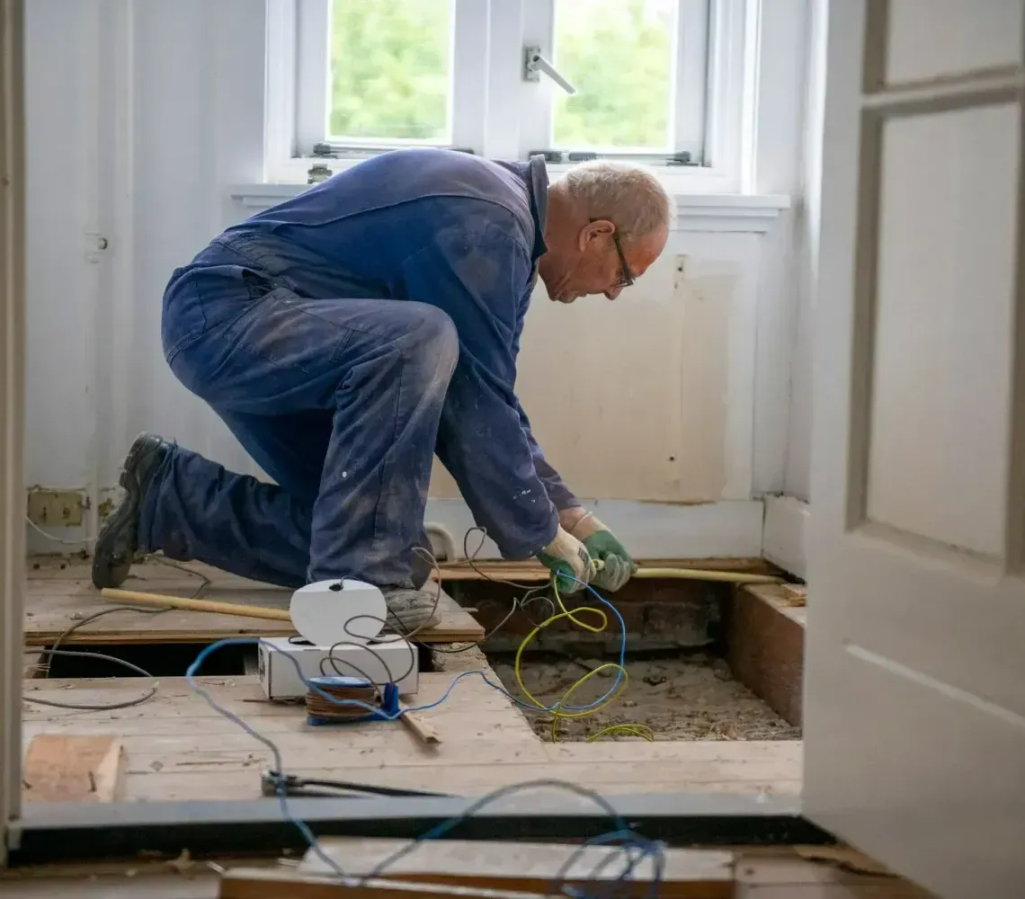 A worker in blue overalls kneels on a wooden floor, reaching into an open space to install electrical wiring.