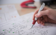 A hand uses a red and silver pen to trace lines on a complex engineering blueprint, with a red stapler in the background.