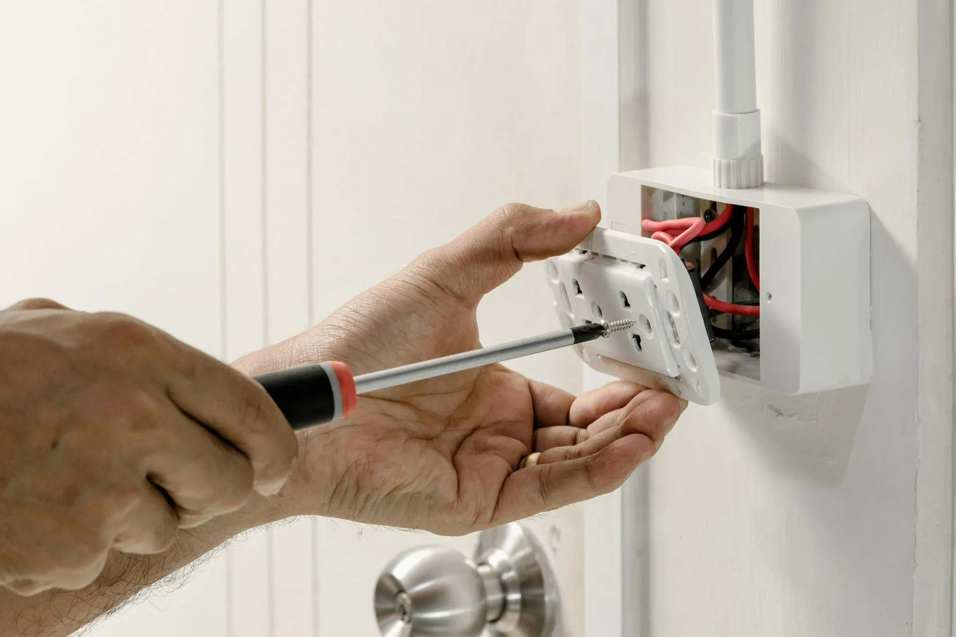 A person uses a screwdriver to attach a white electrical outlet plate to a wall-mounted box with exposed red and black wires.