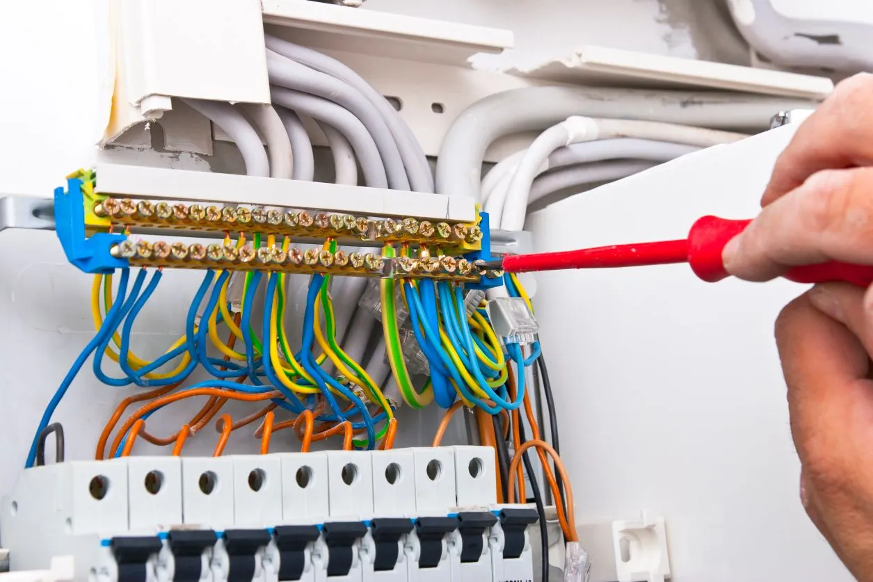 A person uses a red-handled screwdriver to tighten a connection on an electrical terminal block in a circuit breaker panel.
