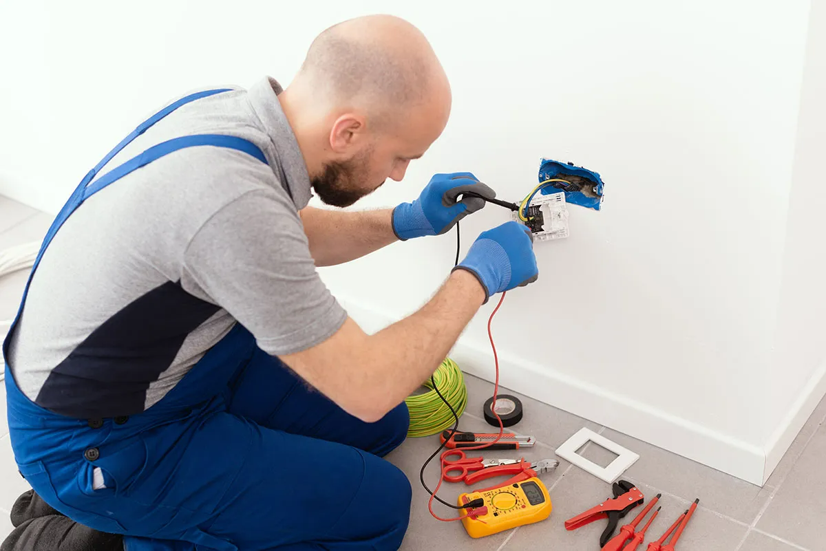 An electrician in blue overalls and gloves works on a wall outlet, with professional tools arranged on the floor.
