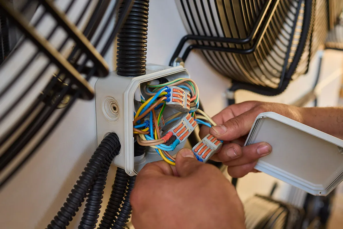 Hands working on electrical wiring inside a wall-mounted junction box located between two black cooling fans.