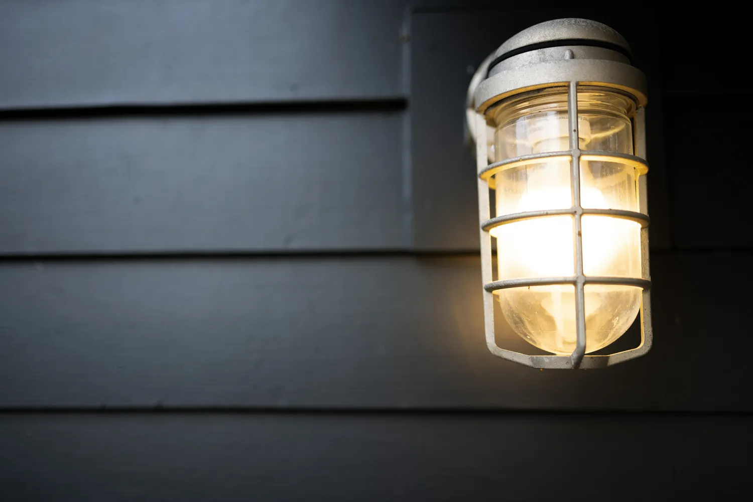 A lit cage-style outdoor wall sconce mounted on dark horizontal siding.