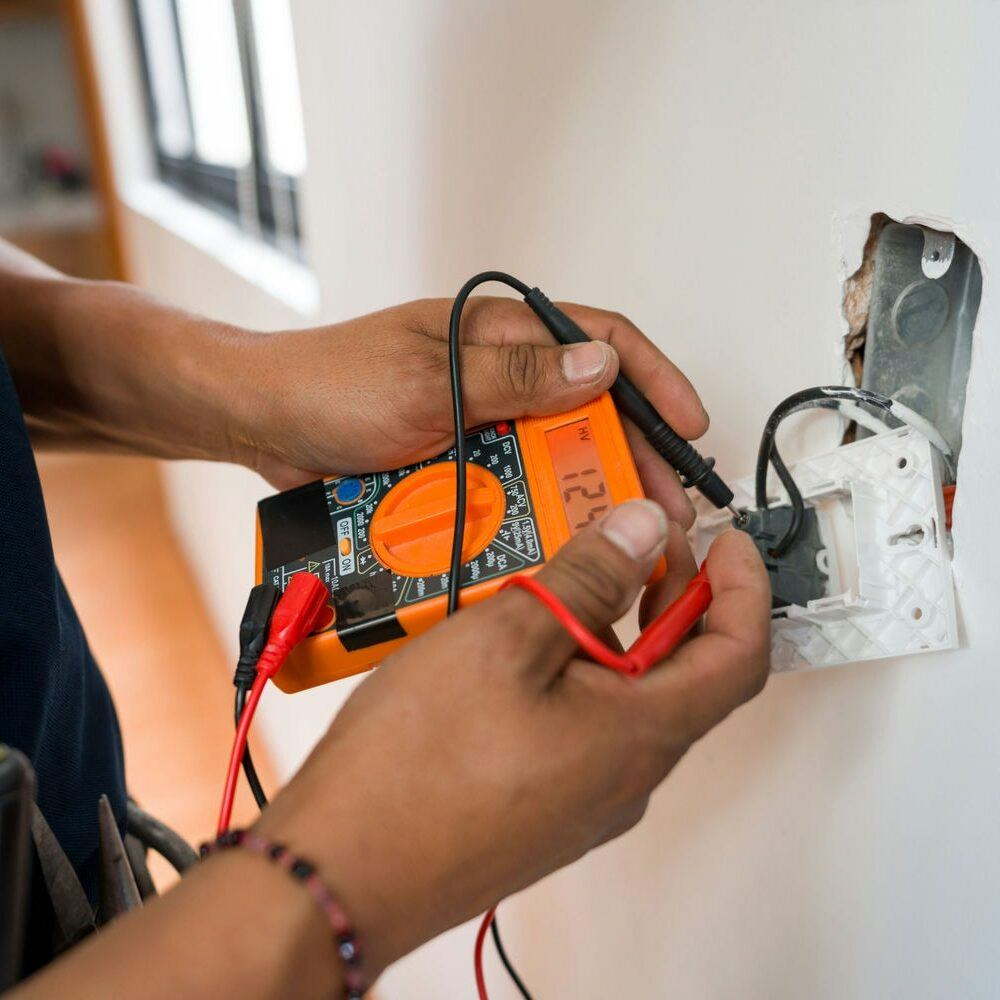 A person uses an orange digital multimeter to test electrical wires inside a wall outlet box.