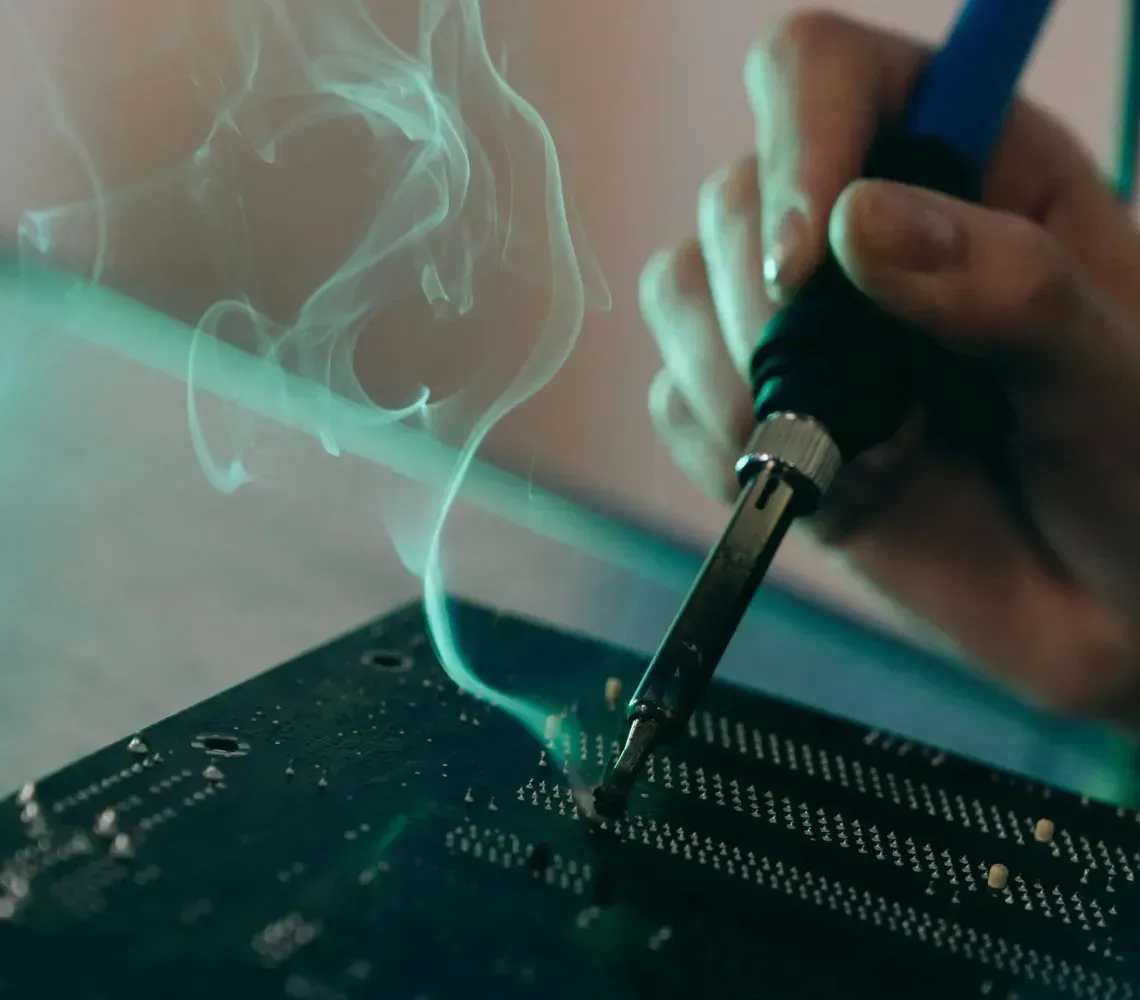 A person uses a soldering iron on a dark circuit board, creating a swirl of smoke.