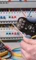 An electrician in gloves uses wire strippers near a circuit breaker panel with a multimeter on the table.