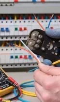 An electrician in gloves uses wire strippers near a circuit breaker panel with a multimeter on the table.