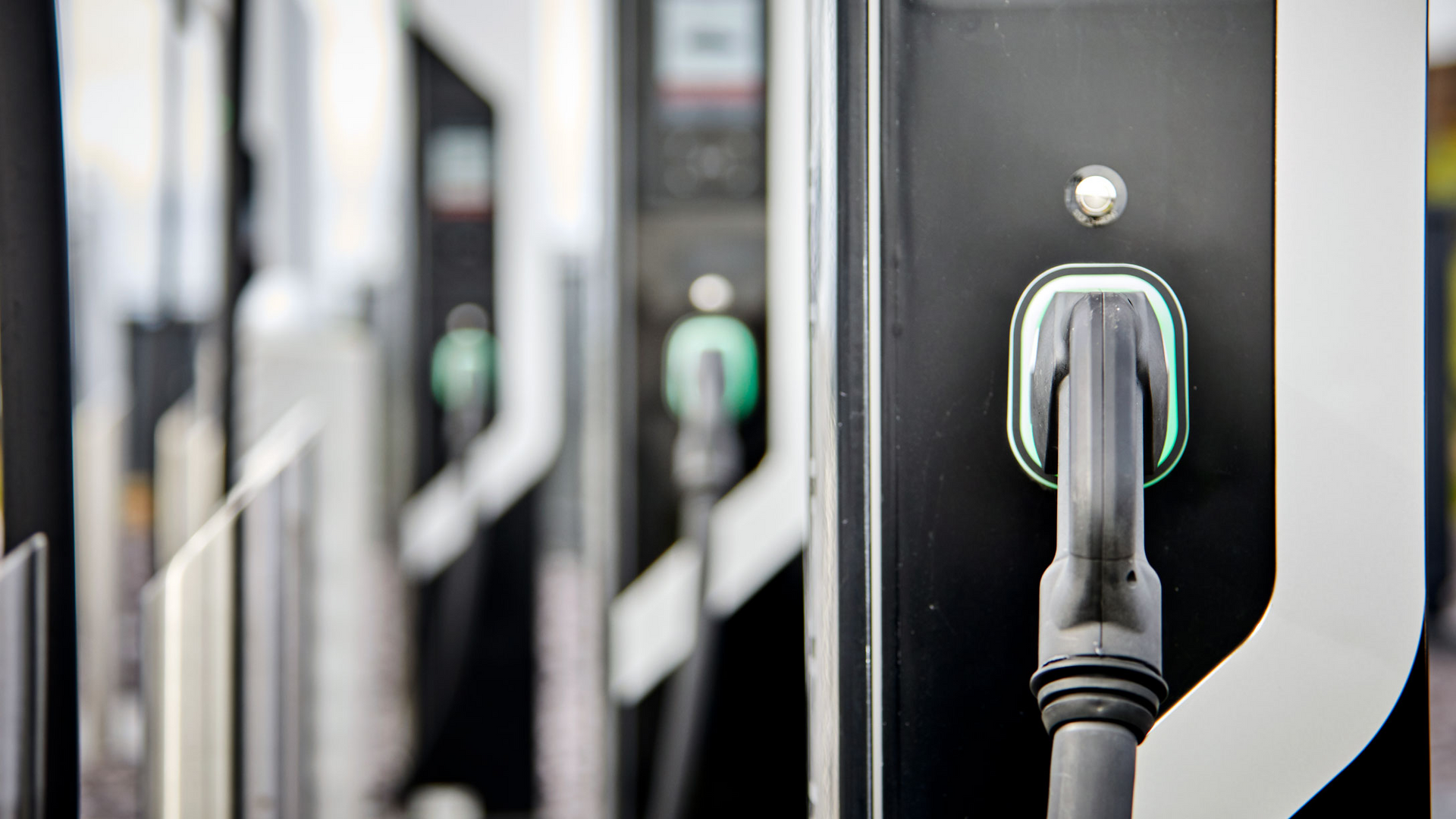 A row of modern electric vehicle charging stations, with a focus on a black charging nozzle docked on a unit.