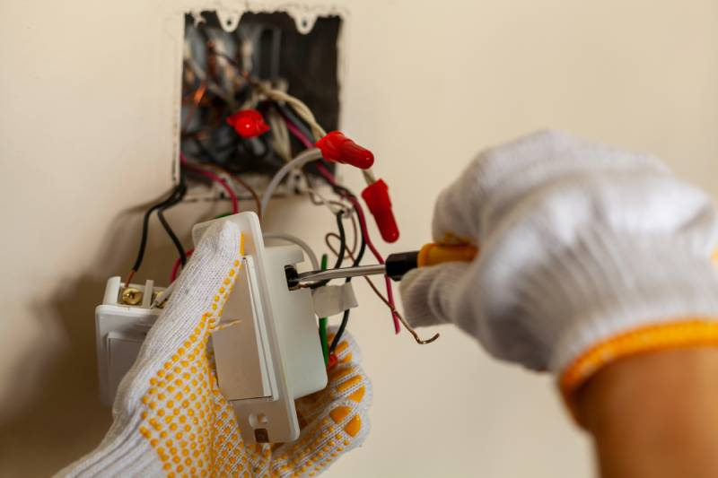 Hands in work gloves use a screwdriver to install a light switch into an electrical wall box.