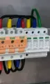 Electrical surge protection device mounted on a rail, featuring orange fuse holders, multicolored wires, and a green ground.