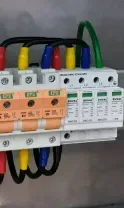 Electrical surge protection device mounted on a rail, featuring orange fuse holders, multicolored wires, and a green ground.
